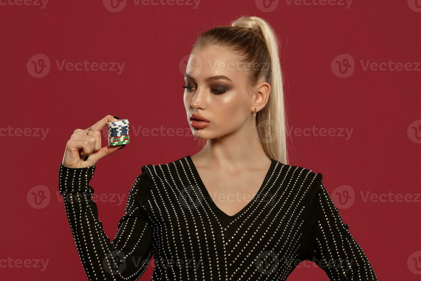 Blonde female with ponytail, in jewelry and black dress in rhinestones. She showing stack of chips, posing on red background. Poker, casino. Close-up photo