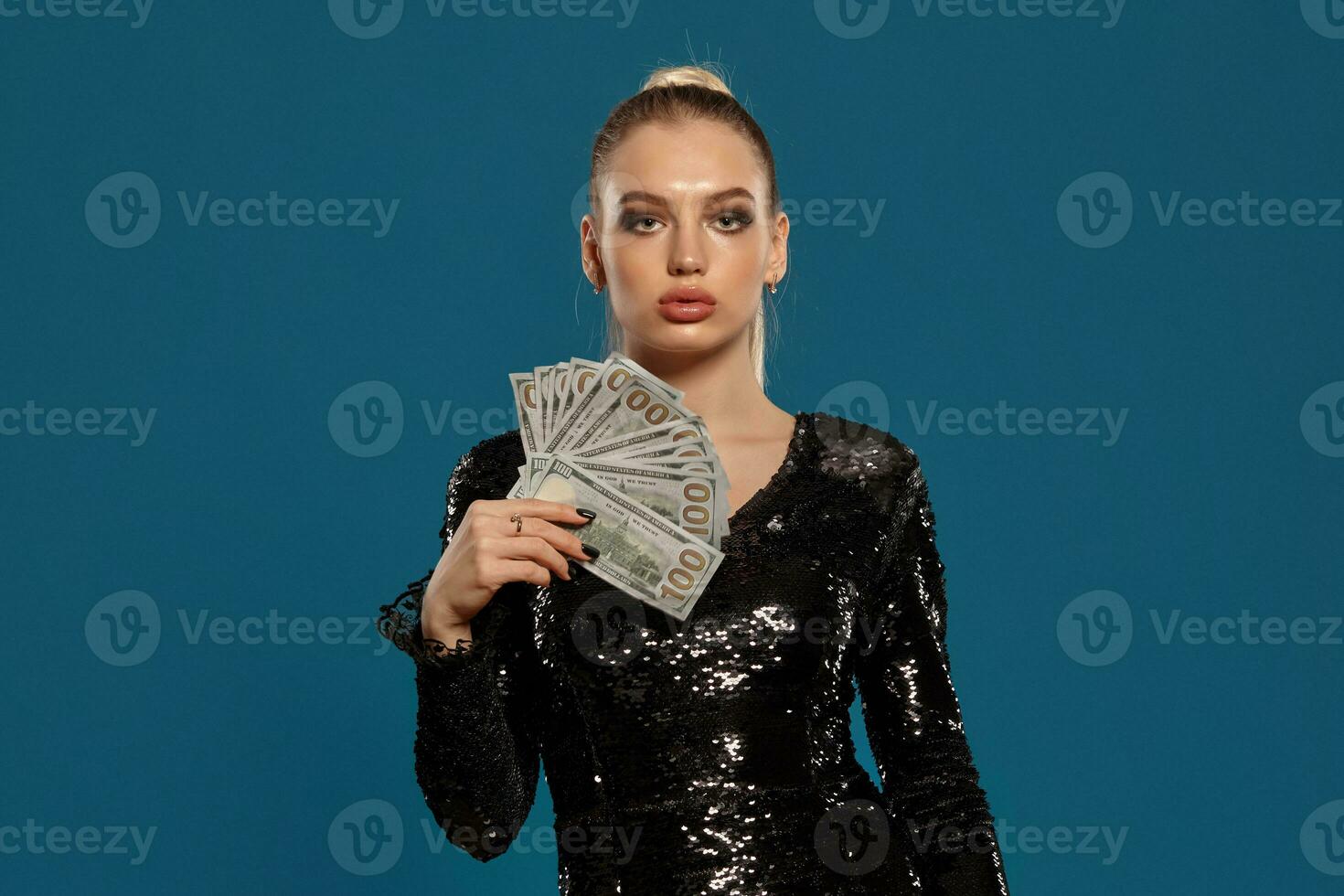 Blonde model with ponytail, in black sequin dress. She is holding some cash, posing on blue background. Gambling, poker, casino. Close-up photo