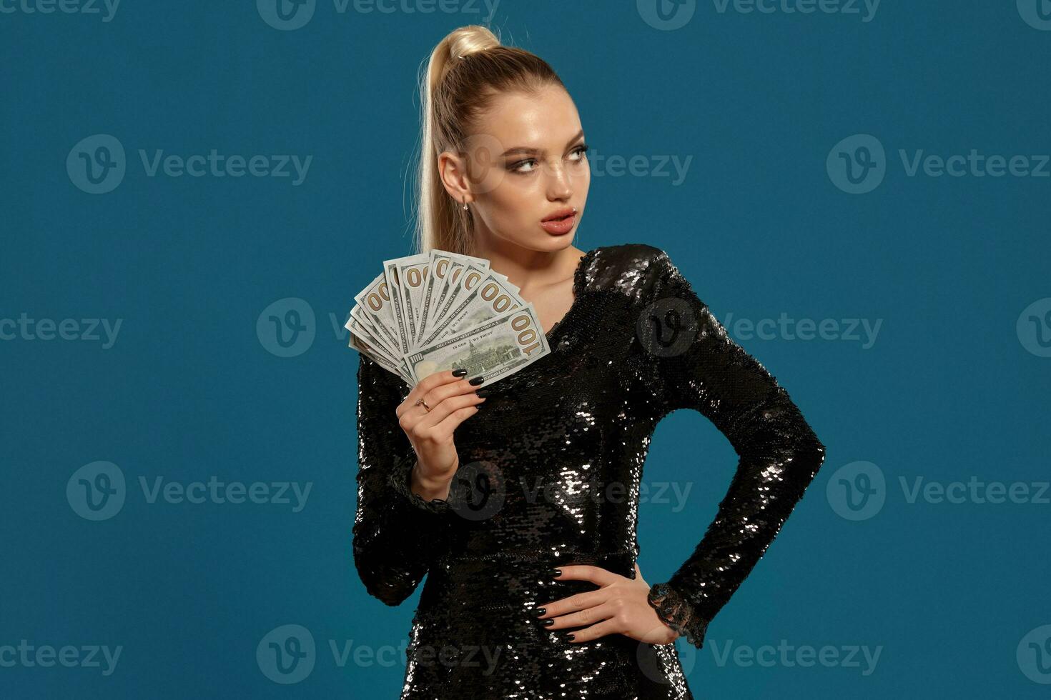 Blonde lady with ponytail, in black sequin dress. Showing fan of hundred dollar bills, posing on blue background. Gambling, poker, casino. Close-up photo