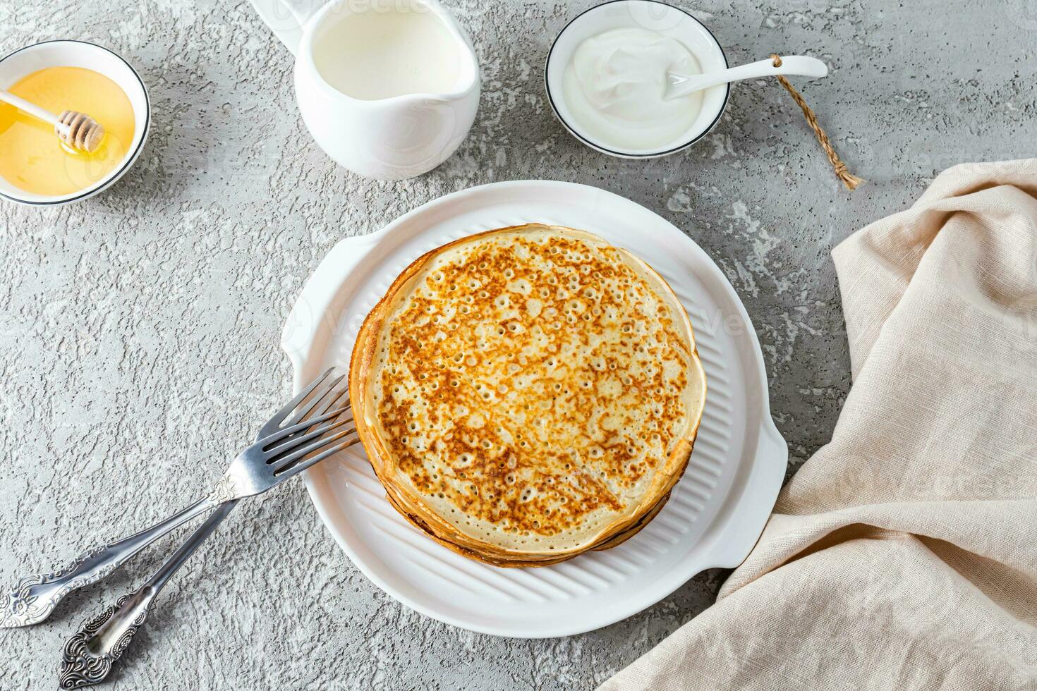 Top view of a stack of delicious pancakes on a white embossed plate, cutlery, sour cream, honey in bowls. Festive traditional breakfast photo