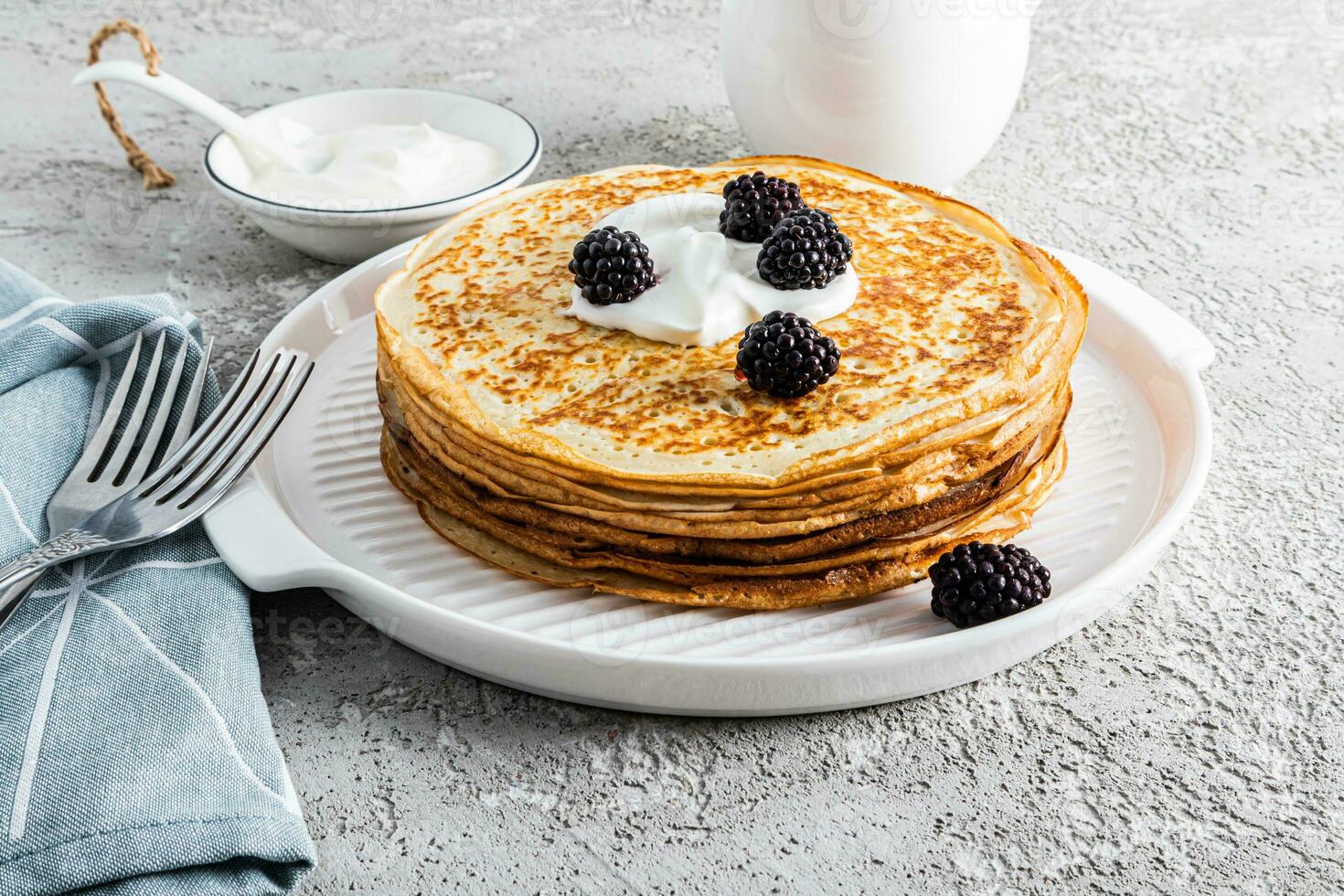 A large stack of delicious hot pancakes with sour cream and blackberries on a beautiful white serving tray with cutlery and a plot. Front view. photo