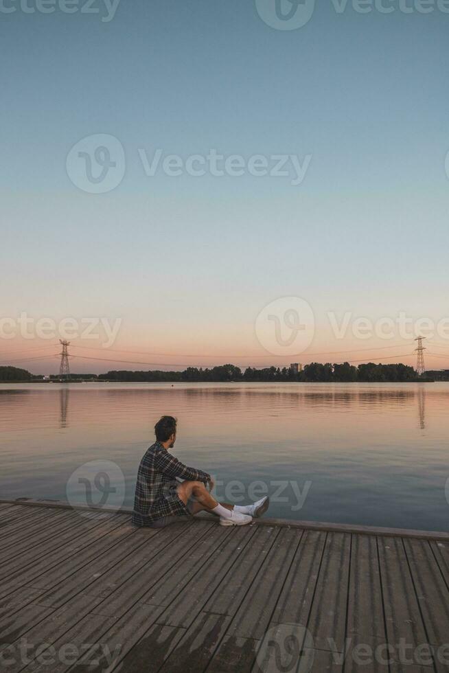 Young 25 year old brown man with plaid shirt sitting on the end of a wooden pier during sunset, watching the calm water surface in Almere Netherlands photo