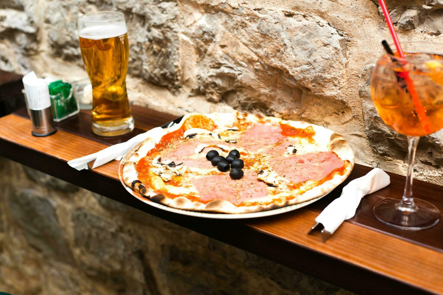 delicioso Pizza con jamón y hongos, un vaso de cerveza y un vaso de aperol a al aire libre cafetería. selectivo enfocar. foto