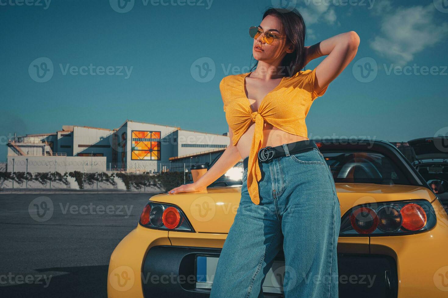 Woman in blue jeans, orange top and sunglasses is posing near yellow car cabriolet with paper cup of coffee on trunk at parking lot. Copy space photo