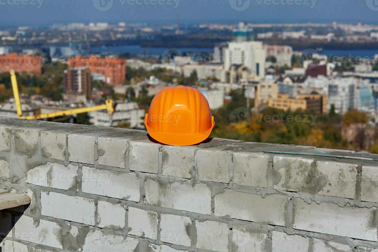 Orange hard hat is on a brick wall. Roof of modern building under construction. Picturesque panoramic cityscape, blue sky, sunny day photo