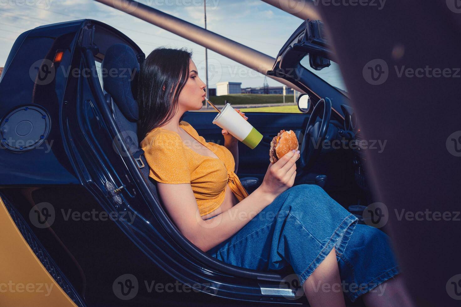 Lady in blue jeans and orange top is drinking beverage from paper cup, holding hamburger while sitting in yellow car cabrio. Fast food. Mock up photo