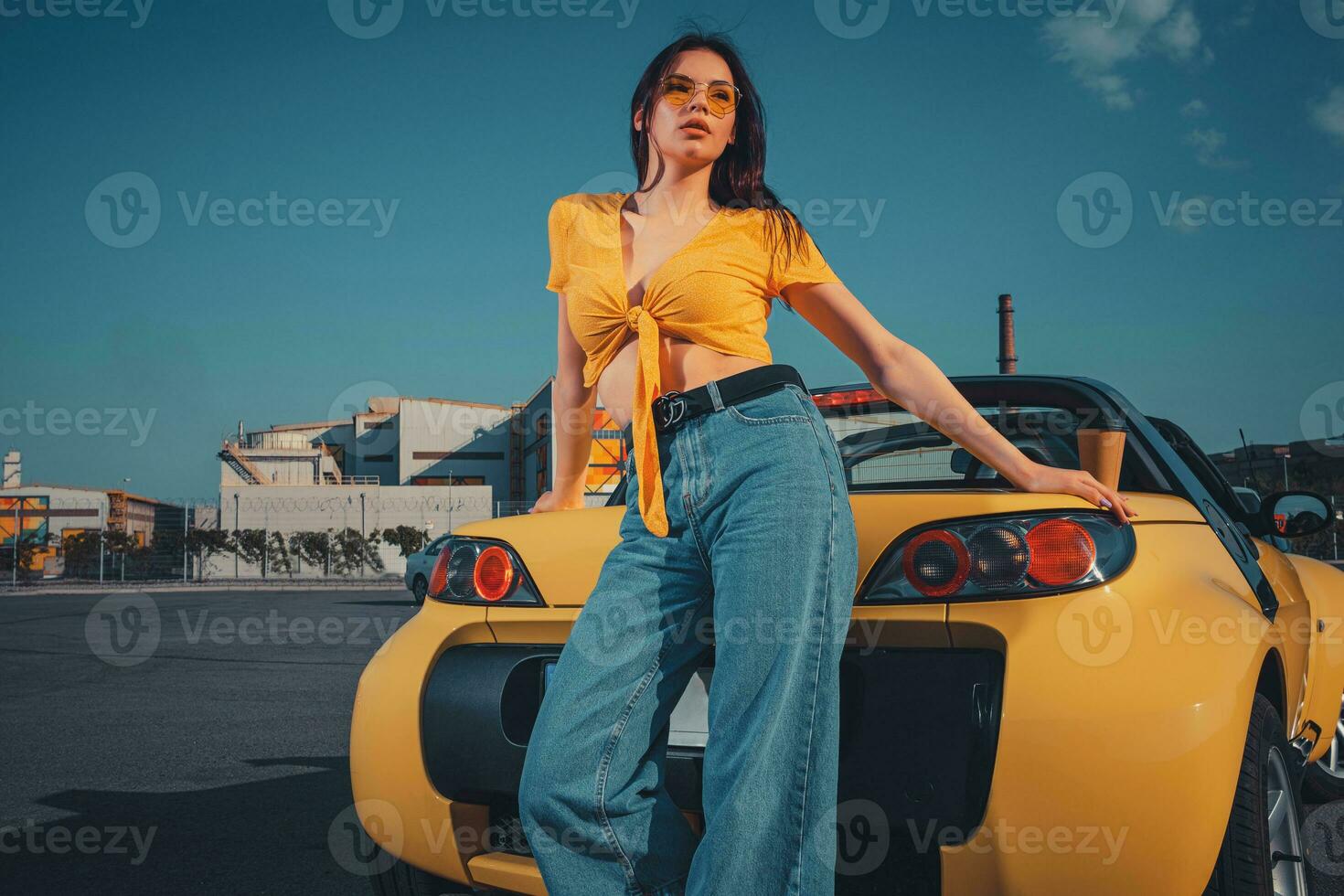 Female in blue jeans, orange top and sunglasses is posing near yellow car roadster with paper cup of tea on trunk at parking lot. Copy space photo