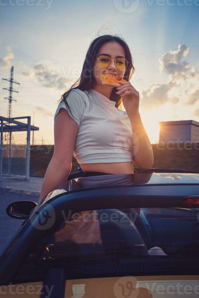 joven hembra en Gafas de sol y blanco camiseta es comiendo Pizza mientras posando en pie en amarillo coche cabriolé. rápido alimento. verano puesta de sol. cerca arriba foto