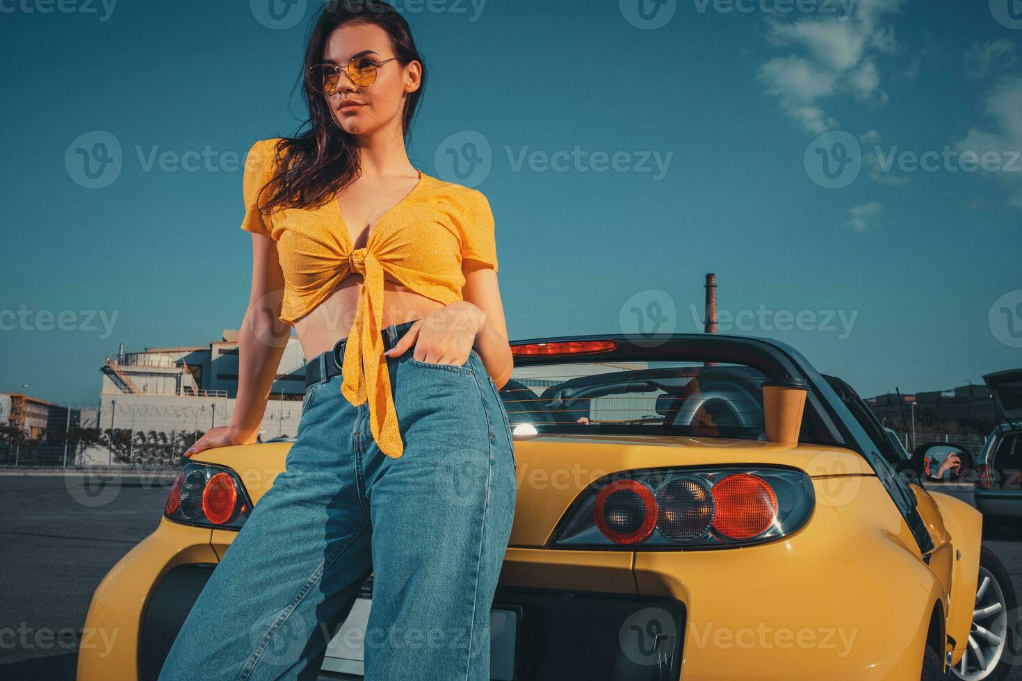 niña en azul vaqueros, naranja parte superior y Gafas de sol es posando propensión en amarillo coche cabriolé con papel taza de bebida en maletero a estacionamiento lote. Copiar espacio foto