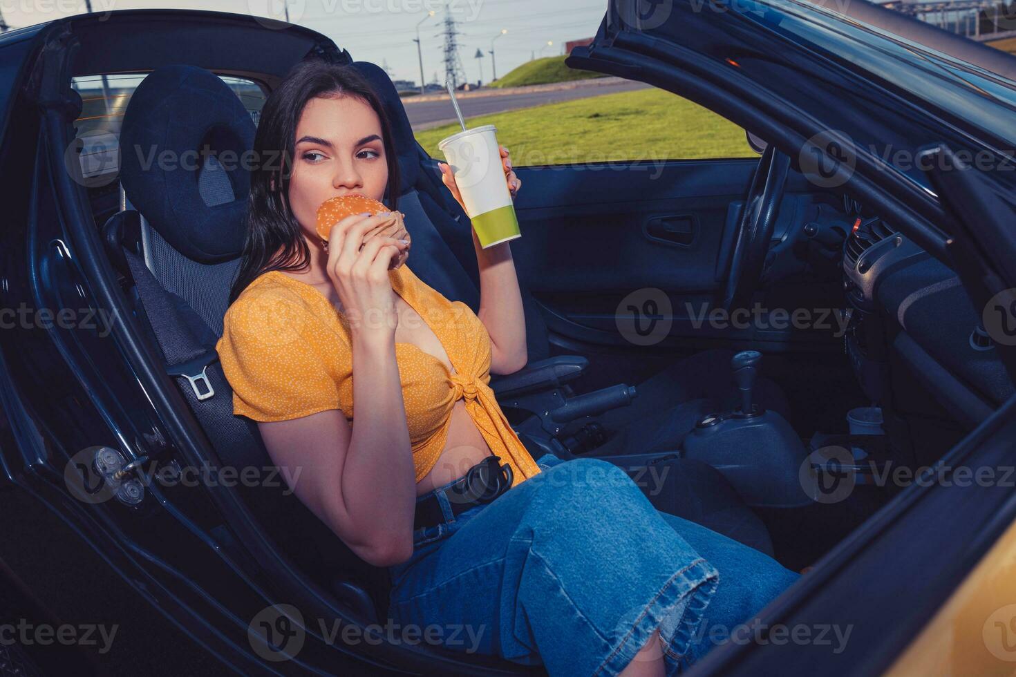 Model in blue jeans and orange top is eating hamburger and holding beverage in paper cup while sitting in yellow car cabriolet. Fast food. Copy space photo