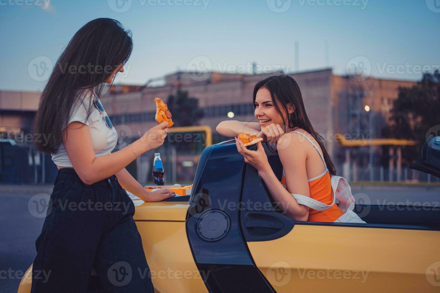 Young girls in casual outfit are smiling, eating pizza, posing in yellow car cabrio with french fries and soda in glass bottle on its trunk. Fast food photo