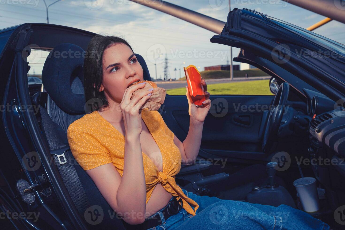 Female in blue jeans and orange top is eating hamburger and holding paper package with french fries while sitting in yellow car. Fast food. Copy space photo