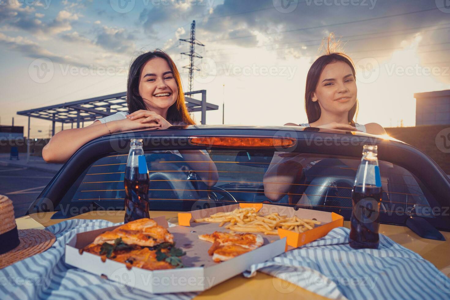 Young women are smiling, posing in yellow car cabriolet with french fries, pizza and soda in glass bottles on its trunk. Fast food. Copy space photo