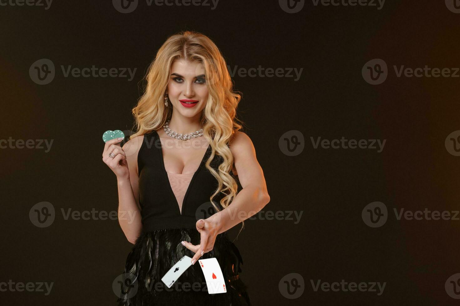 Blonde girl in black dress and necklace. Smiling, showing two green chips, throwing something, posing on brown background. Poker, casino. Close-up photo