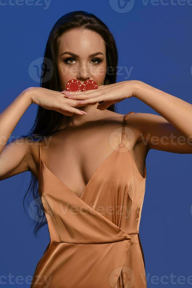 morena modelo en seda beige vestido. ella es participación dos rojo papas fritas, posando en contra azul estudio antecedentes. juego, póker, casino. de cerca foto