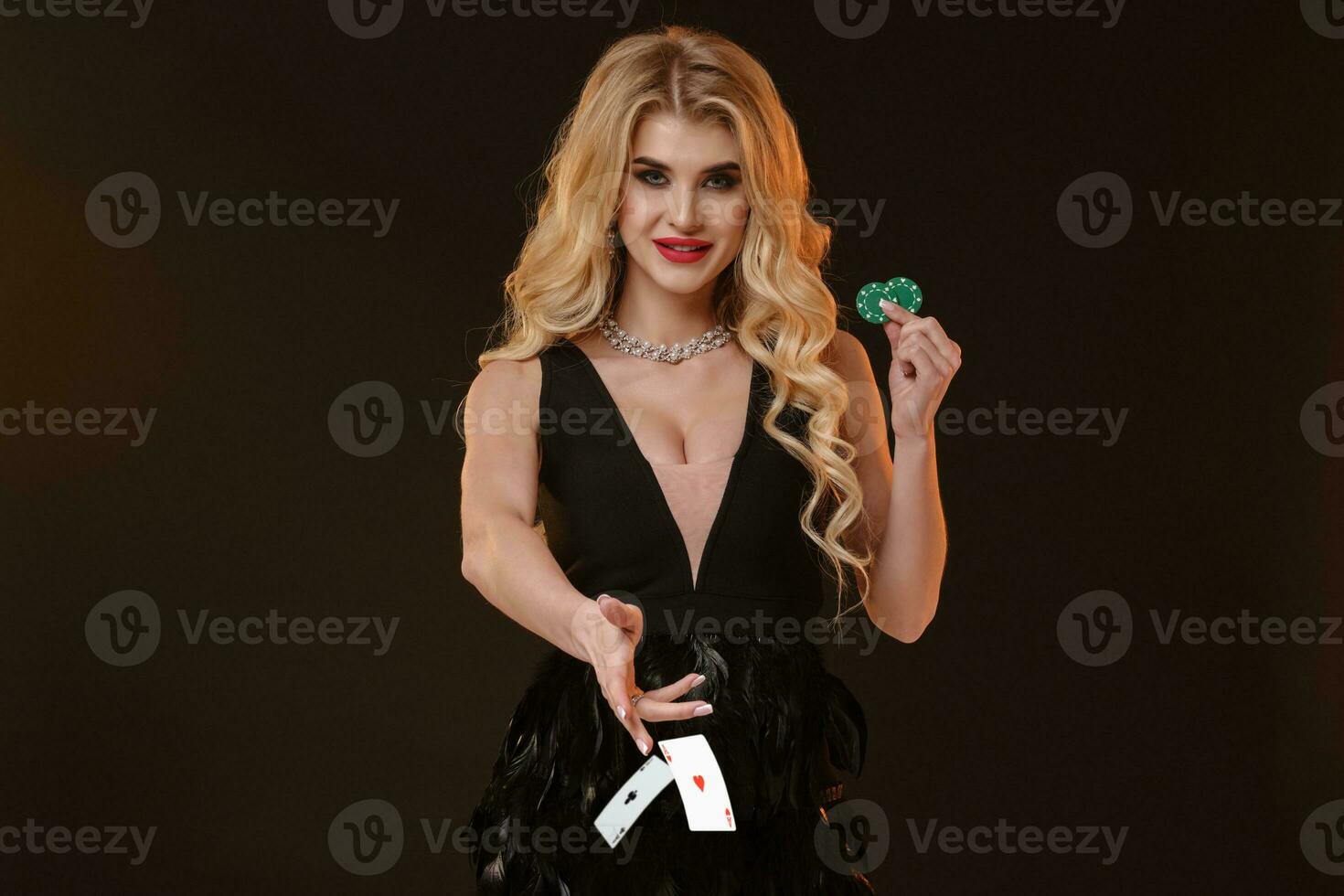 rubia modelo en negro vestir y collar. sonriente, demostración dos verde papas fritas, lanzamiento algo, posando en marrón antecedentes. póker, casino. de cerca foto