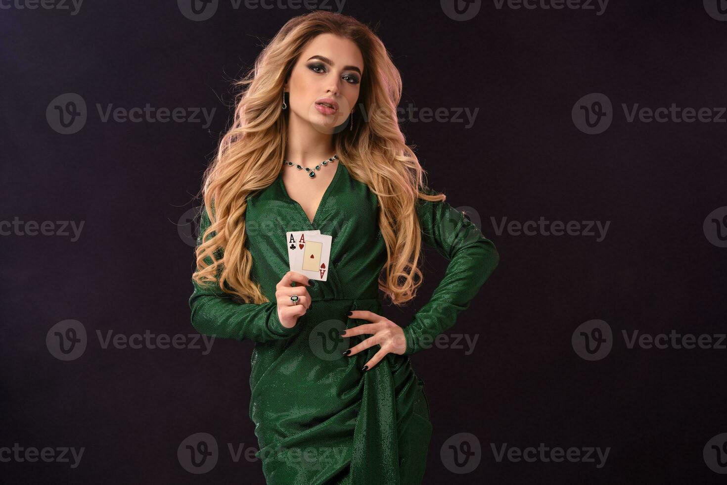 Blonde lady in green stylish dress and jewelry. Put her hand on hip, showing two playing cards, posing on black background. Poker, casino. Close-up photo