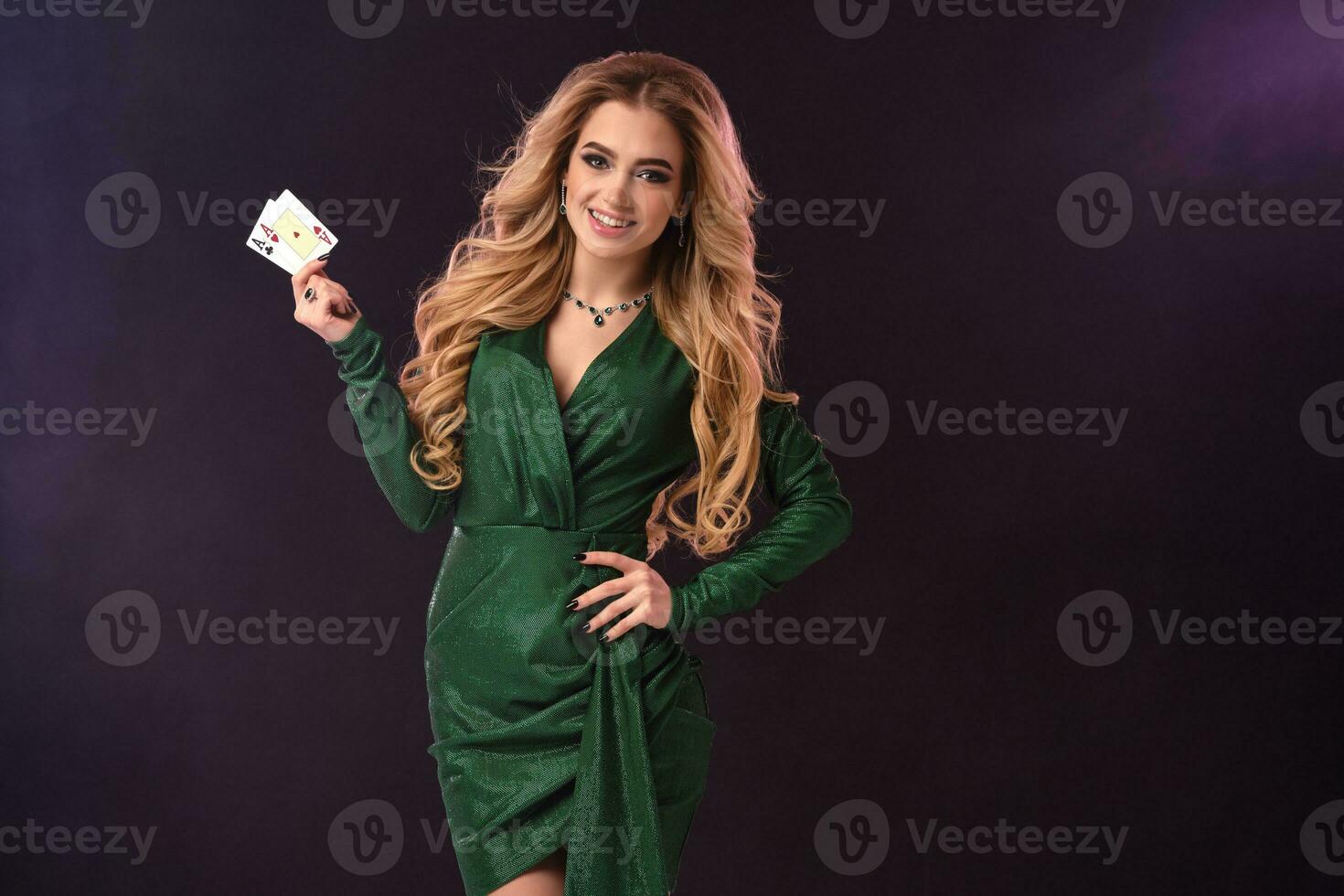 rubia mujer en verde elegante vestir y joyas. poner mano en cadera, sonriente, demostración dos ases, posando en negro antecedentes. póker, casino. de cerca foto