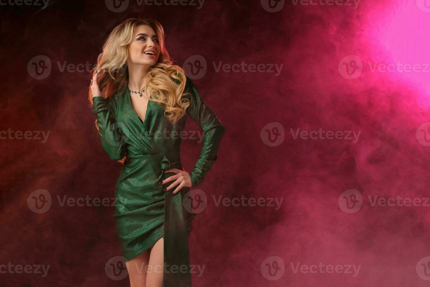 maravilloso rubia Rizado modelo en verde elegante vestir y joyas. ella tiene poner su mano en cintura, sonriente, posando en vistoso ahumado antecedentes. cerca arriba foto