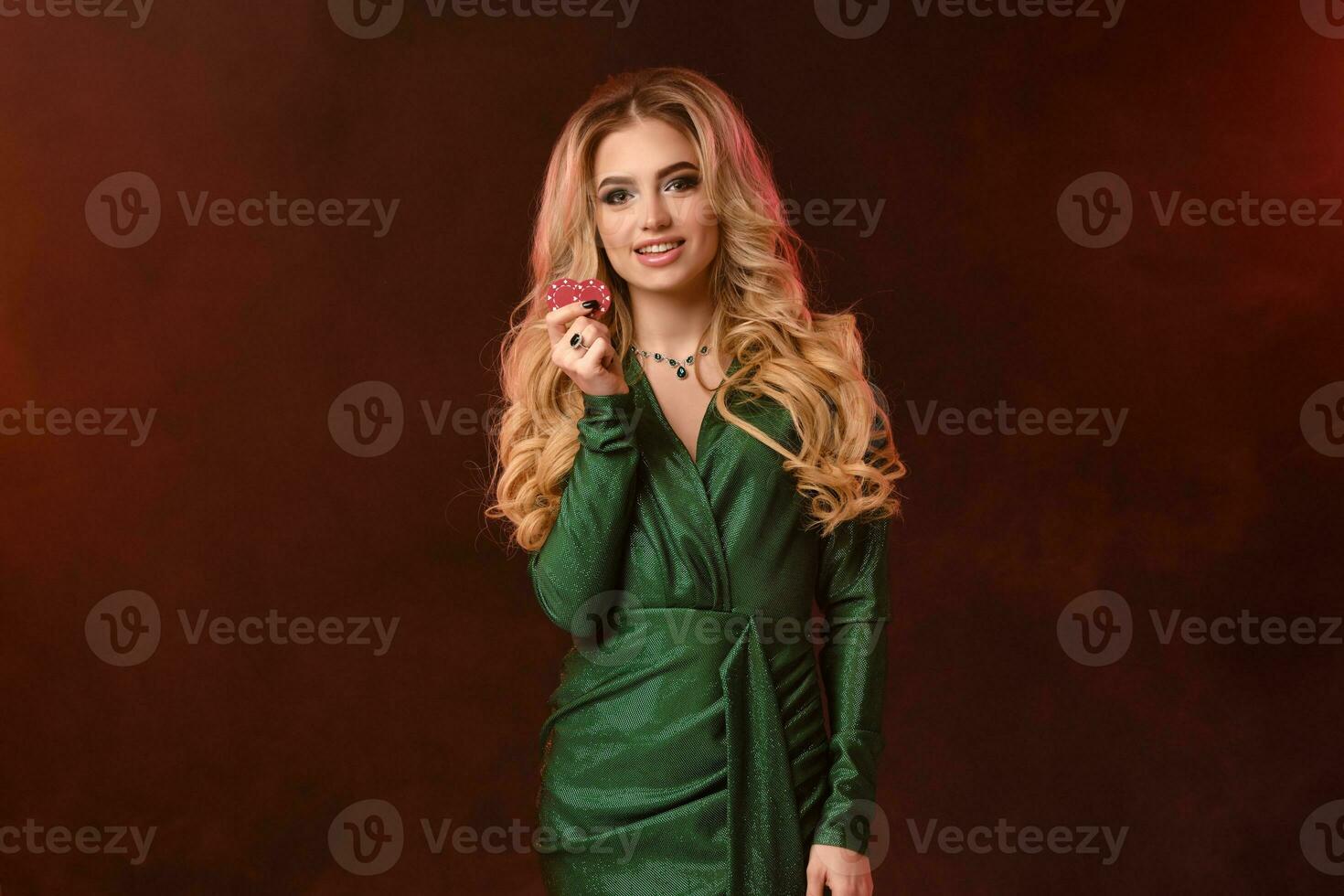Blonde curly model, bright make-up, in green stylish dress and jewelry. Smiling, showing two red chips, posing on brown smoky background. Close-up photo