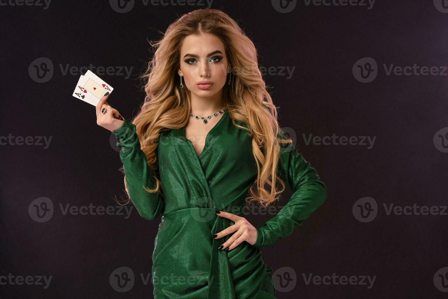 rubia niña con constituir, en verde elegante vestir y joyas. poner mano en cadera, demostración dos ases, posando en negro antecedentes. póker, casino. de cerca foto