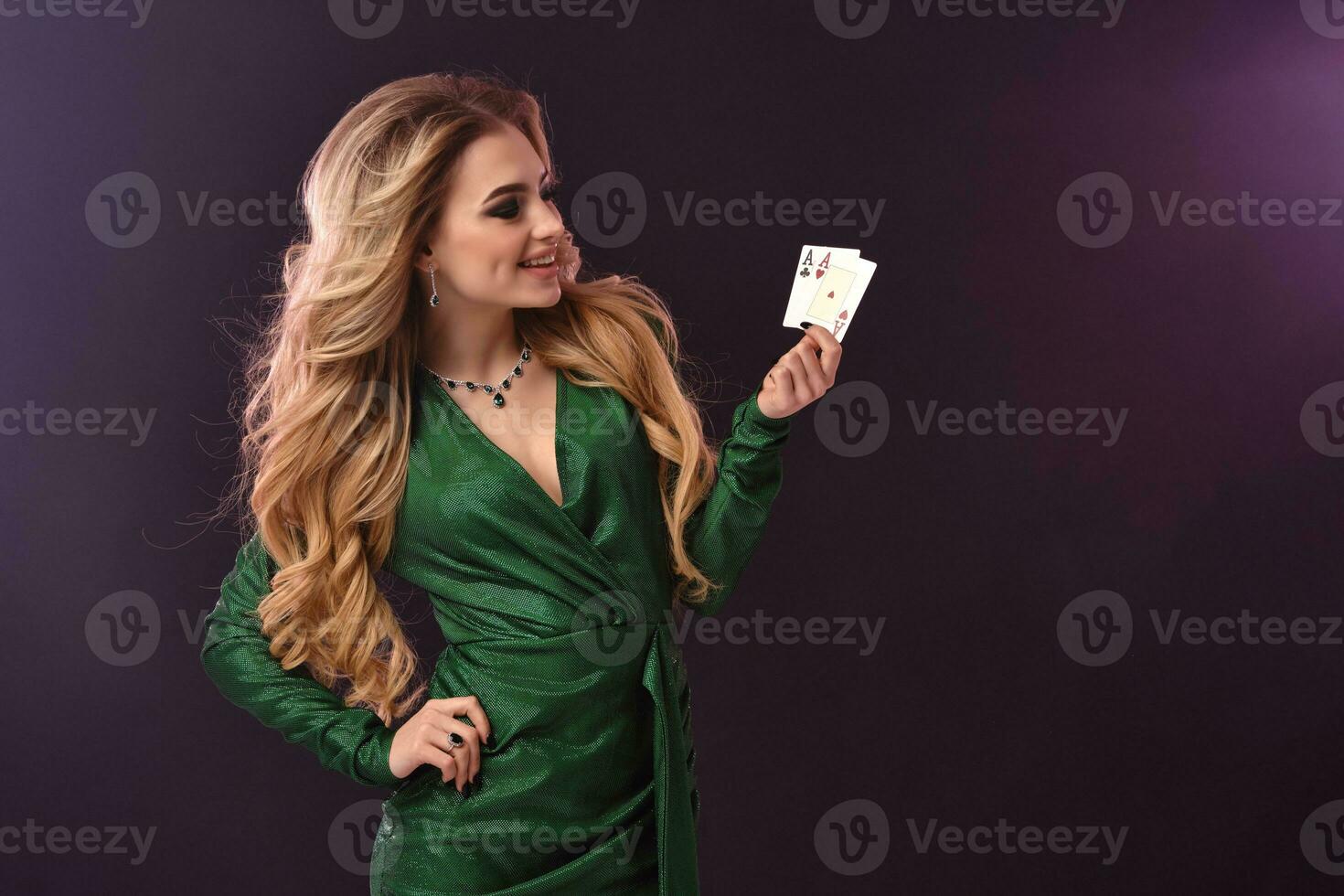 Blonde female in green stylish dress and jewelry. Put hand on hip, smiling, showing two aces, posing on purple background. Poker, casino. Close-up photo