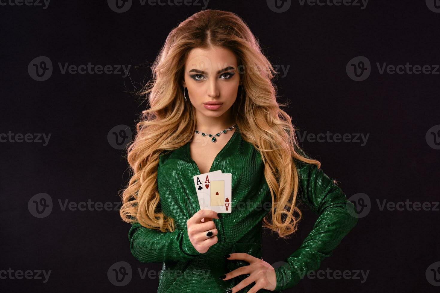 Blonde model in green stylish dress and jewelry. Put her hand on hip, showing two playing cards, posing on black background. Poker, casino. Close-up photo