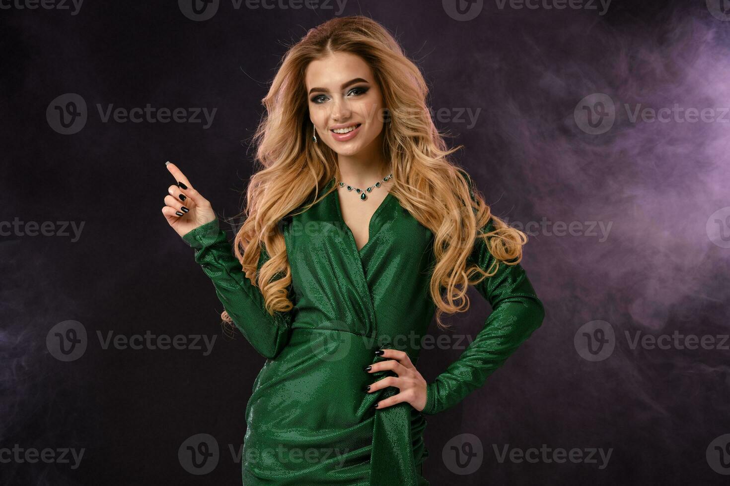 Blonde girl in green stylish dress and jewelry. Smiling, pointing at something by forefinger, posing on black smoky background. Close up, copy space photo