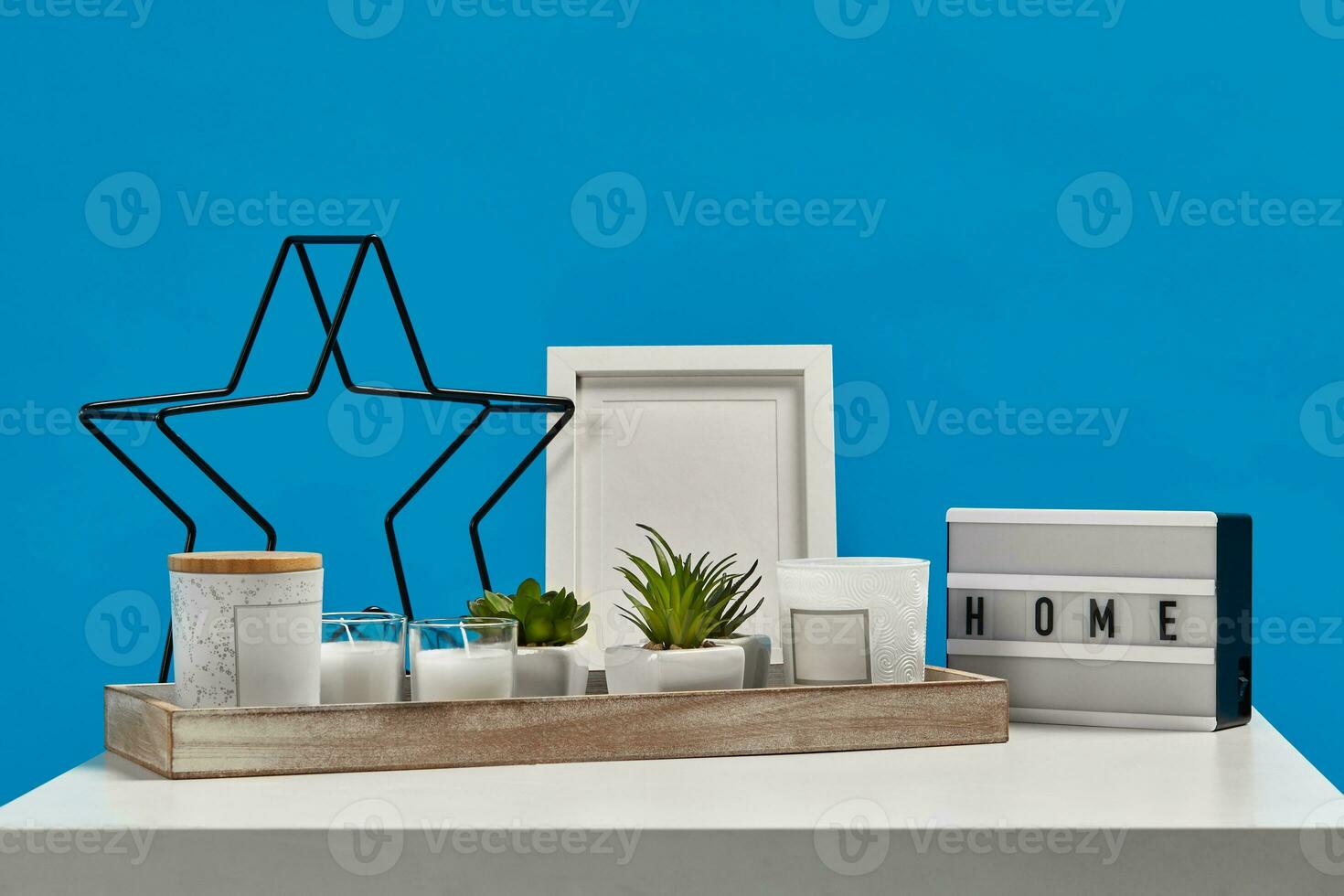 White table, wooden stand with green succulents, candles, jars. Black iron star, lamp inscription home, empty photo frame. Blue background. Close up