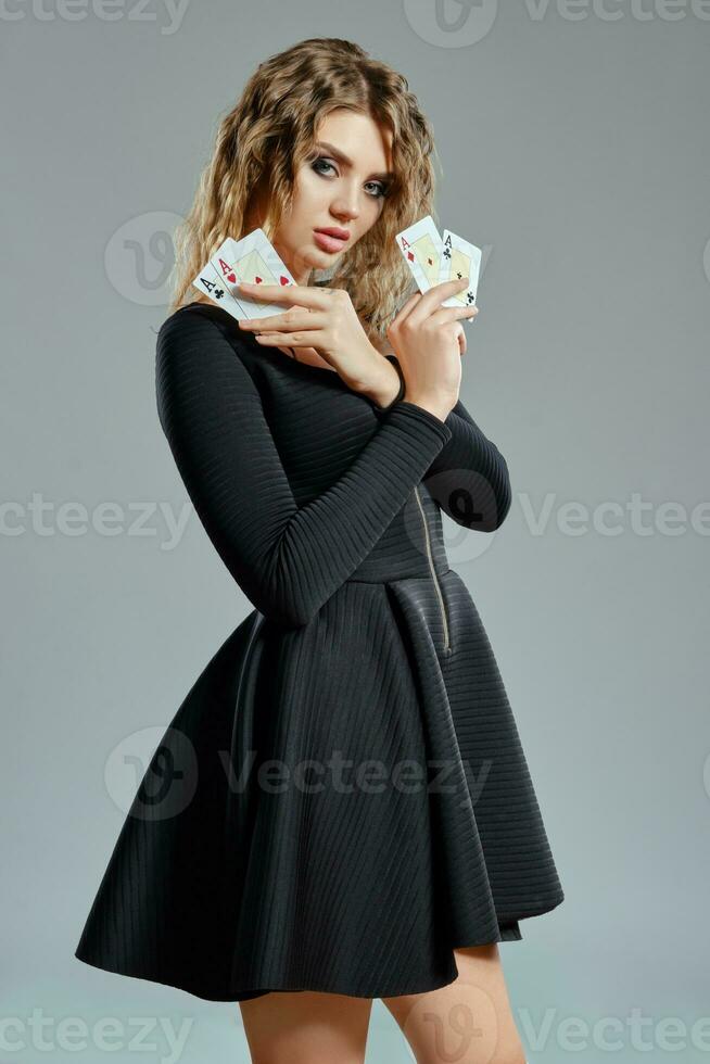 Blonde maiden with curly hair, in black short dress showing four aces, posing on gray background. Gambling entertainment, poker, casino. Close-up. photo