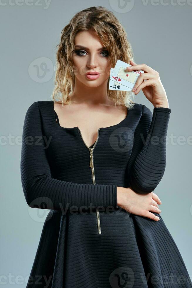Blonde girl in black dress showing two aces, posing against gray background. Gambling entertainment, poker, casino. Close-up. photo