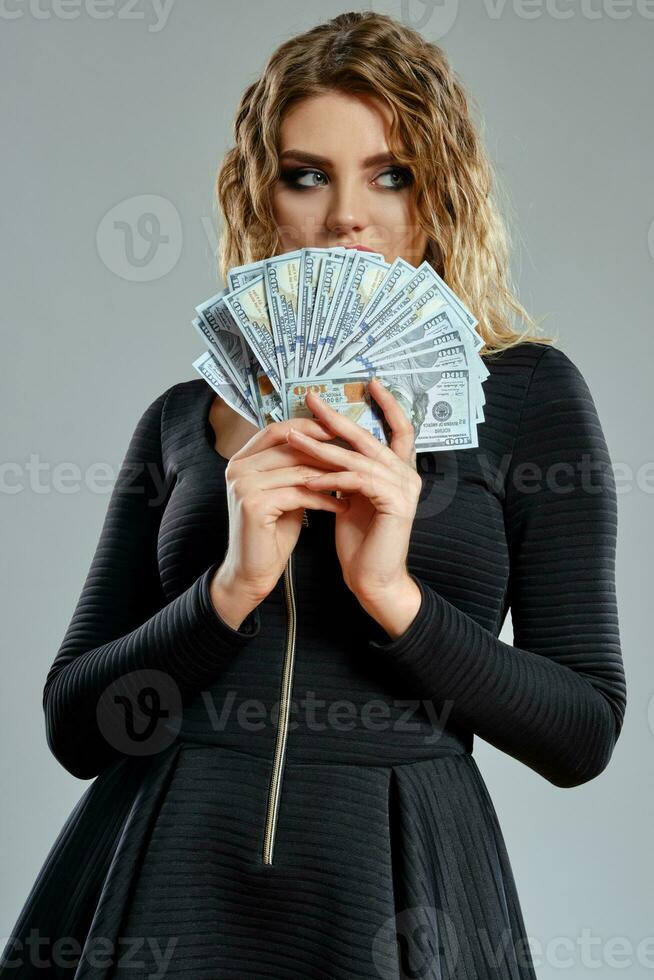Blonde lady in black short dress is holding some cash, posing against gray background. Gambling entertainment, poker, casino. Close-up. photo