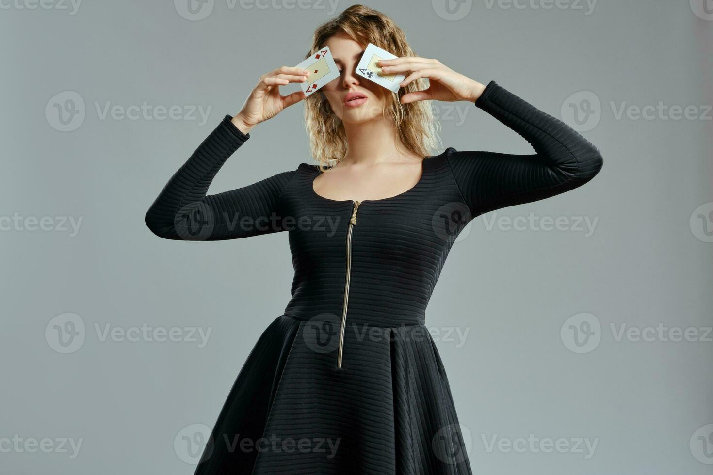 Blonde lady in black dress has covered her eyes with two playing cards, posing on gray background. Gambling entertainment, poker, casino. Close-up. photo