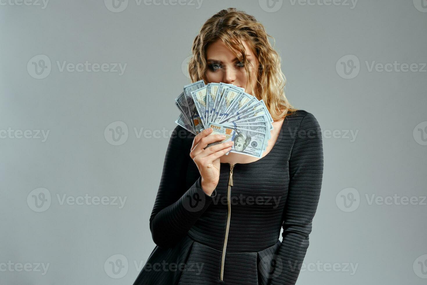 Blonde woman in black dress is holding some cash, posing against gray background. Gambling entertainment, poker, casino. Close-up. photo