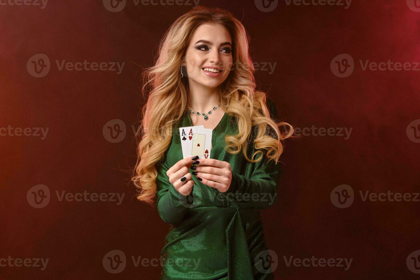 Blonde lady in green stylish dress and jewelry. Smiling and showing two aces, posing on colorful smoky background. Poker, casino. Close-up photo