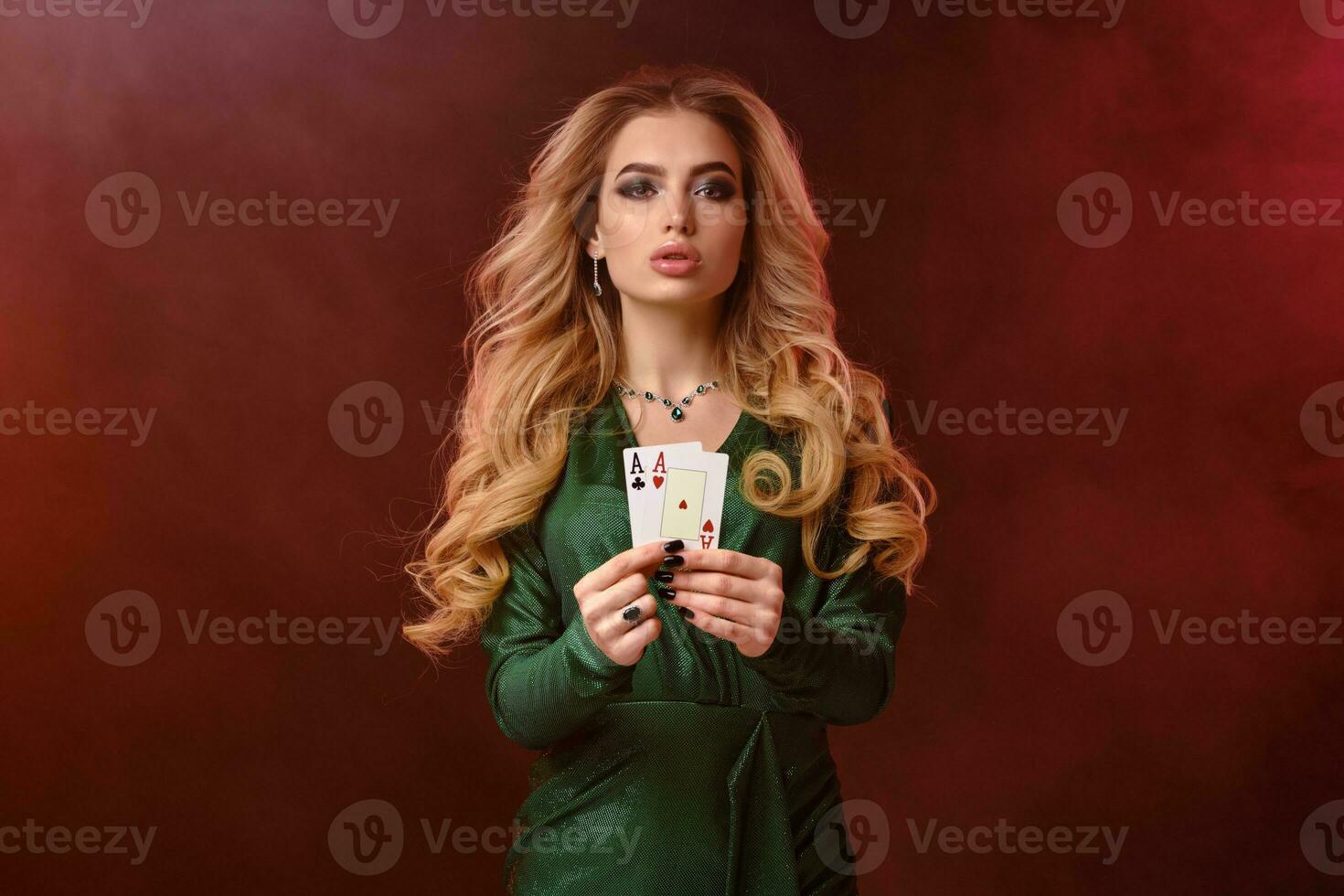 Curly blonde girl in green stylish dress and jewelry. Showing two playing cards, posing on colorful smoky background. Poker, casino. Close-up photo