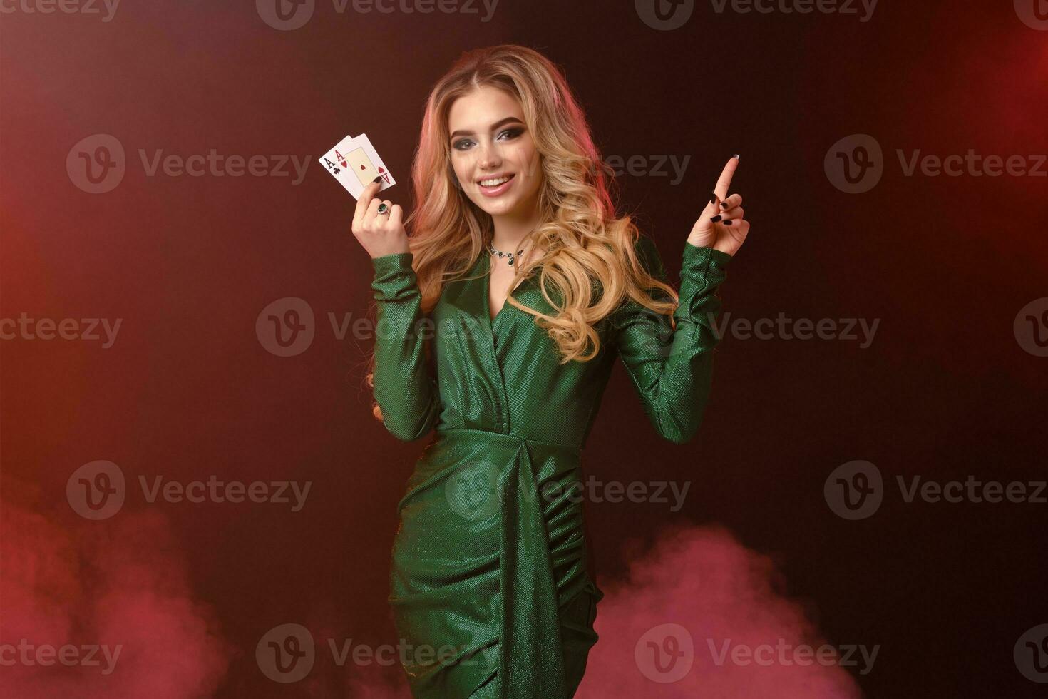 Blonde female in green dress, jewelry. Smiling, showing two aces, pointing at something, posing on colorful smoky background. Poker, casino. Close-up photo