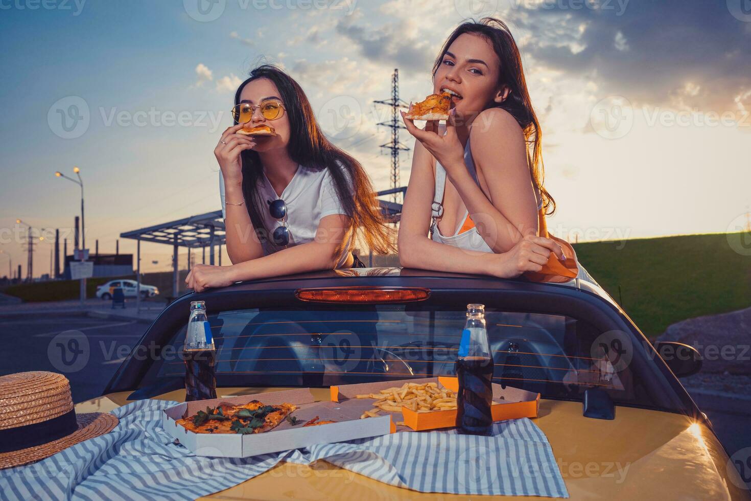 Charming females are eating pizza while posing in yellow car cabriolet with french fries and soda in glass bottles on trunk. Fast food. Close up photo