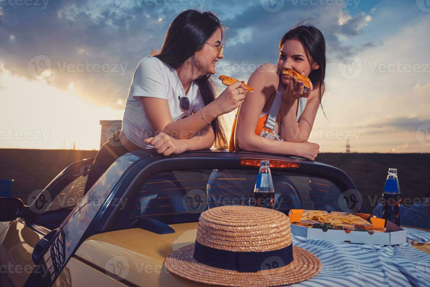 Two young girls are eating pizza, laughing, posing in yellow car with french fries, hat and soda in glass bottles on trunk. Fast food. Mock up photo