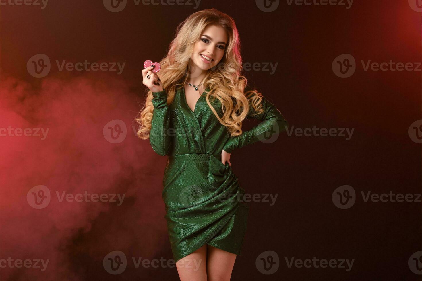 Gorgeous blonde lady in green stylish dress and jewelry. Smiling, showing two red chips, hand on waist, posing on colorful smoky background. Close-up photo