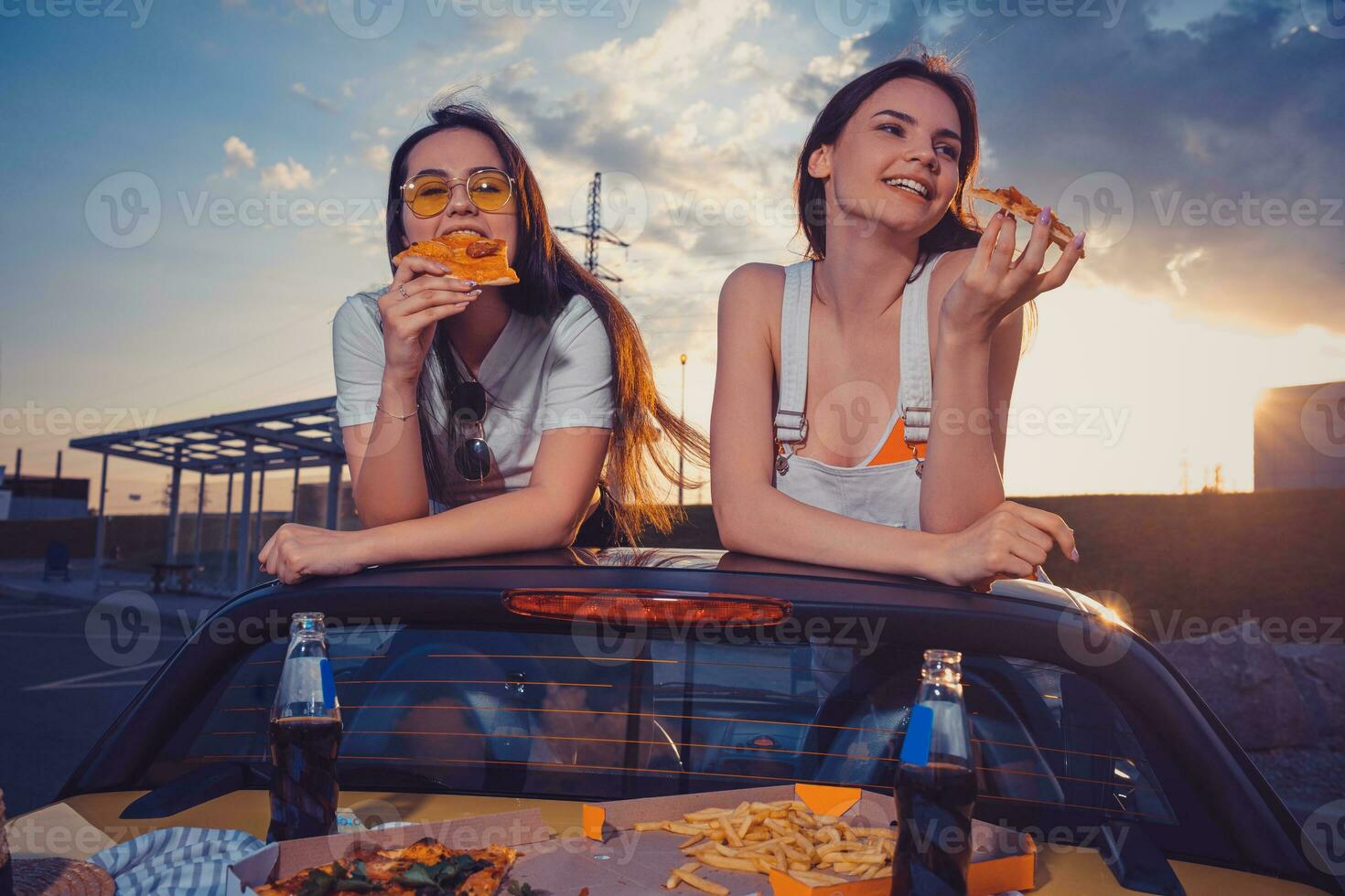 Charming women are eating pizza and smiling while posing in yellow car with french fries and soda in glass bottles on trunk. Fast food. Copy space photo