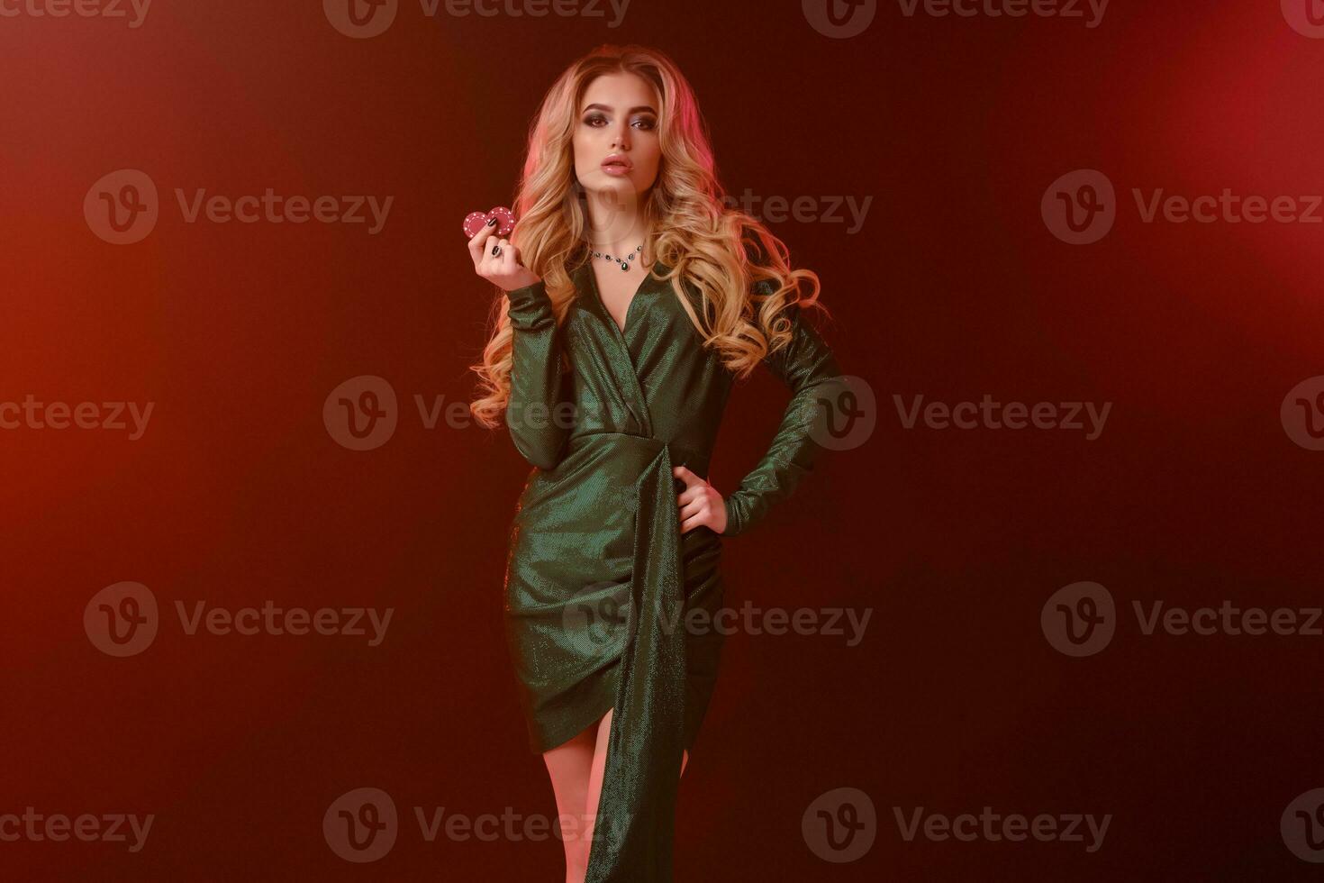 Blonde female with bright make-up, in green stylish dress and jewelry. Showing two red chips, posing on colorful background with backlight. Close-up photo