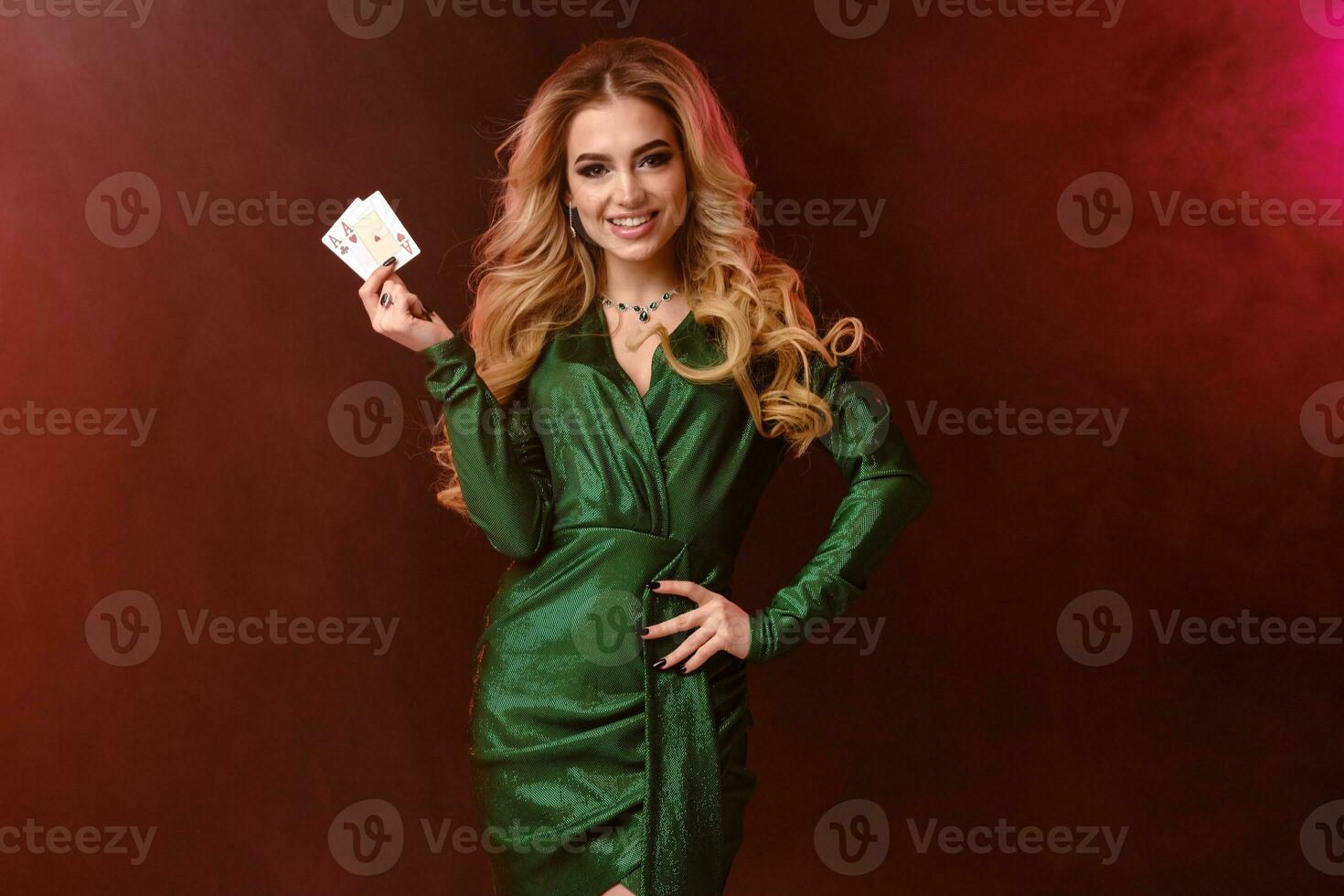 rubia modelo en verde elegante vestir y joyas. sonriente, mano en cadera, demostración dos ases, posando en vistoso ahumado antecedentes. póker, casino. de cerca foto