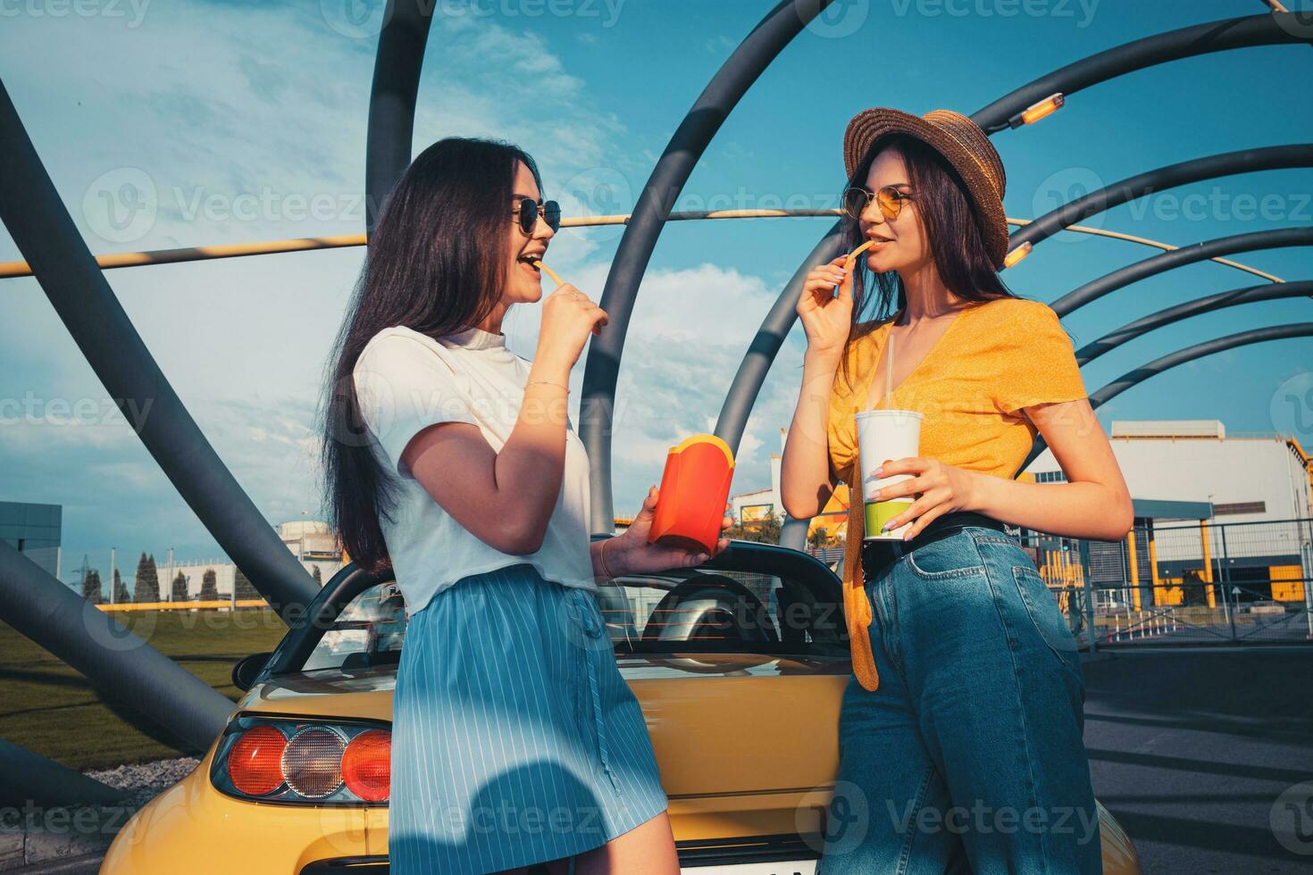 Young females in casual outfit are smiling, eating french fries and holding beverage in paper cup while posing near yellow car. Fast food photo