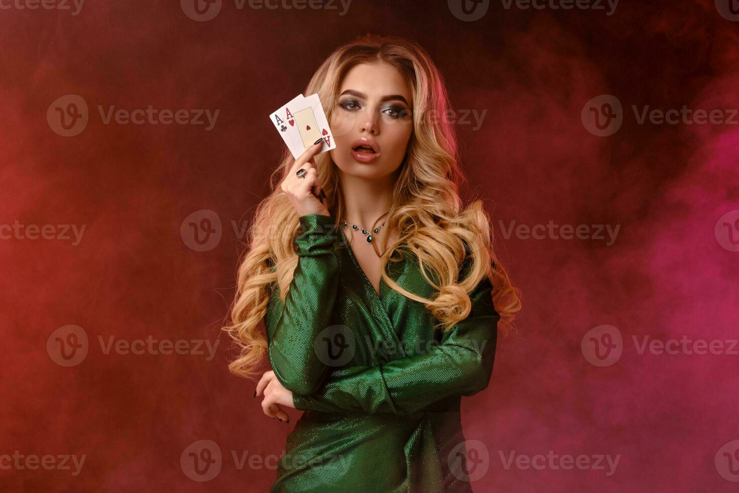 Blonde woman in green dress and jewelry. Showing two playing cards, opened her mouth, posing on colorful smoky background. Poker, casino. Close-up photo