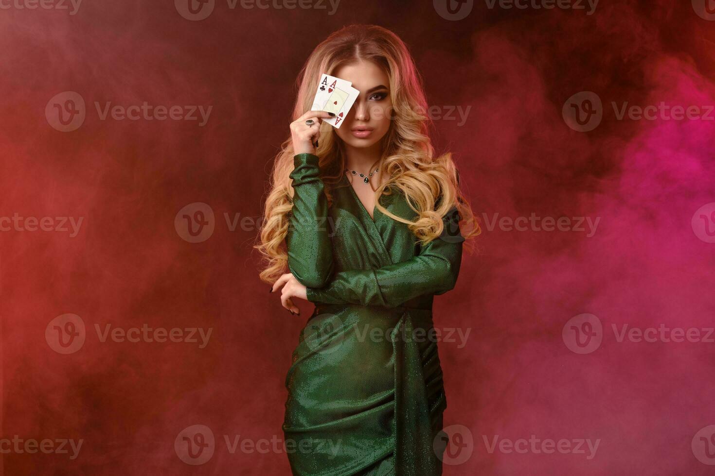 Blonde model in green stylish dress and jewelry. Covered her eye with two playing cards, posing on colorful smoky background. Poker, casino. Close-up photo