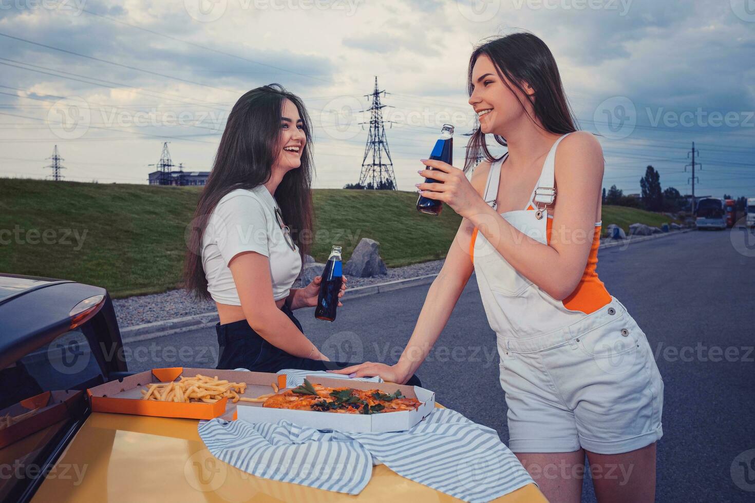 joven muchachas sonriente, disfrutando soda en vaso botellas, posando propensión en maletero de amarillo coche con Pizza y francés papas fritas en él. rápido alimento. Copiar espacio foto