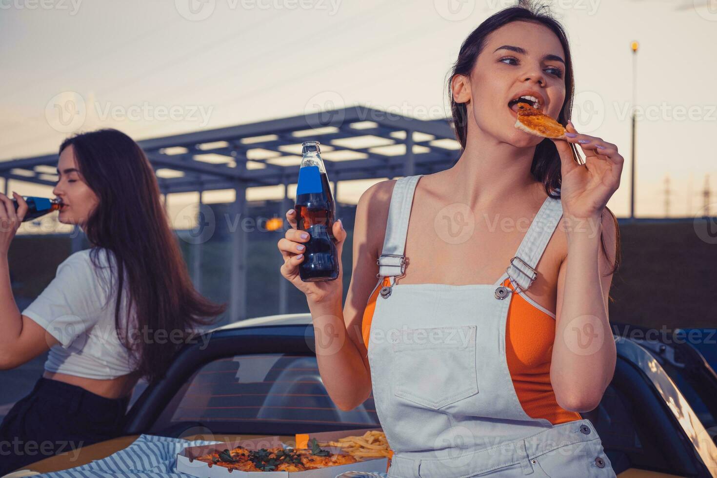 atractivo muchachas en casual atuendo comiendo pizza, disfrutando gaseado bebida en vaso botellas mientras posando cerca amarillo coche en estacionamiento lote. Copiar espacio foto