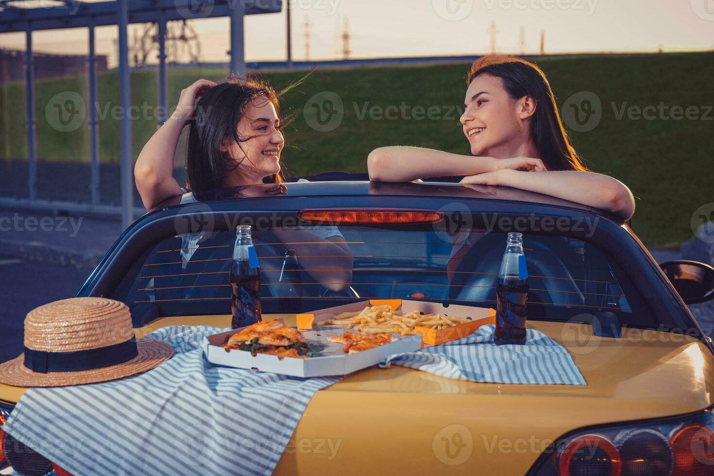 Pretty models are smiling, posing in yellow car with french fries, pizza, hat and soda in glass bottles on its trunk. Fast food. Close up, copy space photo