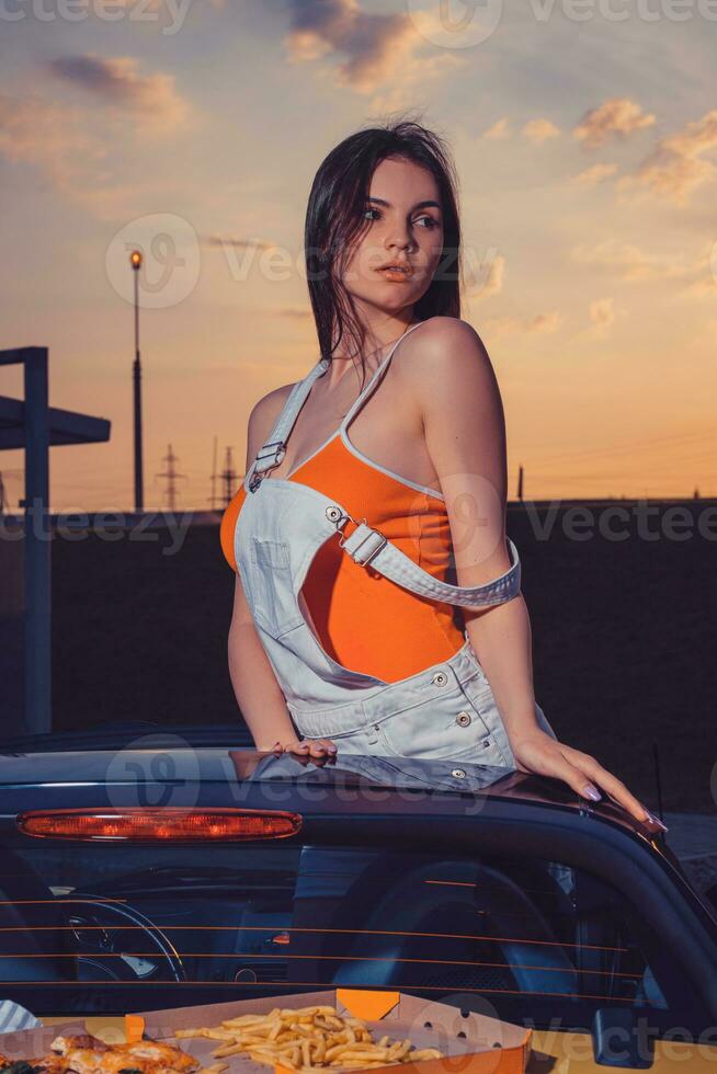 Sexy woman in white overall and orange t-shirt is posing standing in yellow car cabrio with french fries and pizza on its trunk. Fast food. Close up photo